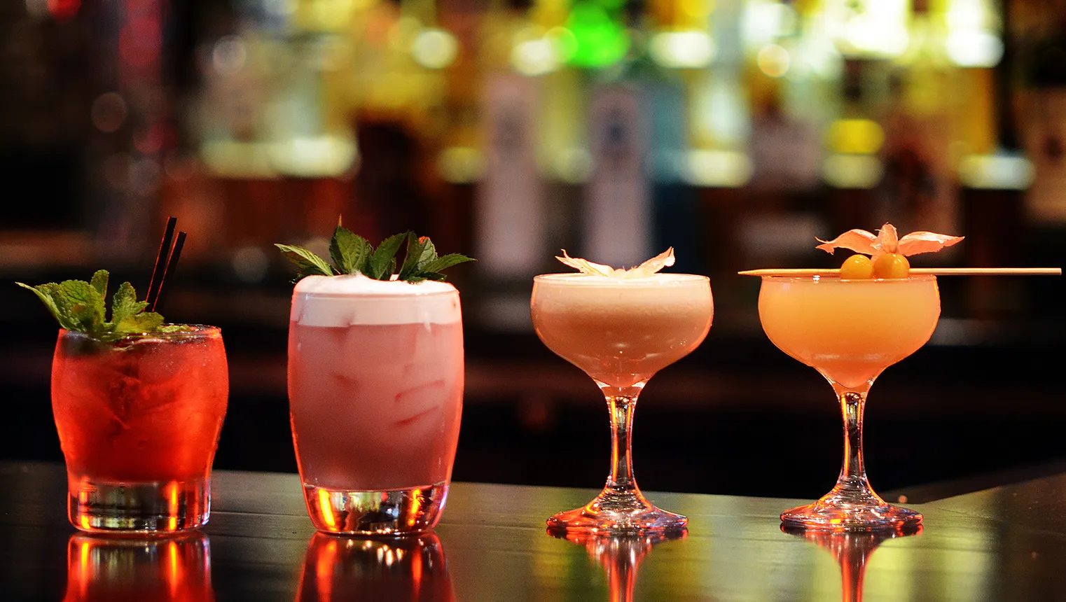 An image of four cocktails in the foreground of different colors and a blurred bar in the background.