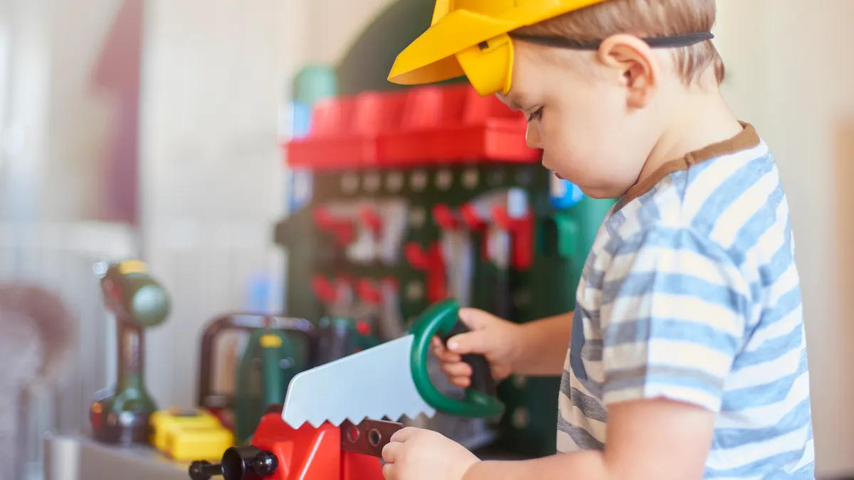 A toddler is wearing a hard hat and using a toy saw at a pretend workbench.