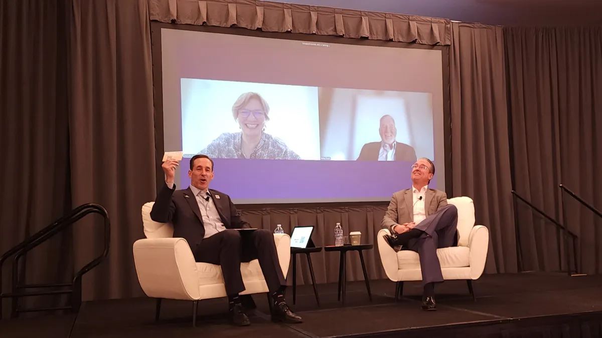 Two people in suits sitting on chairs on stage, and two people on the screen behind them.