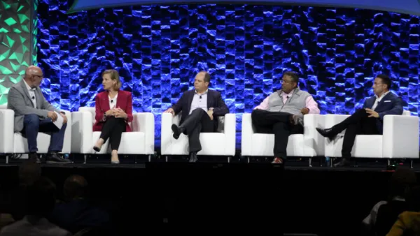 Five people sit in white chairs on a stage. One of them, the second from the left, is speaking.