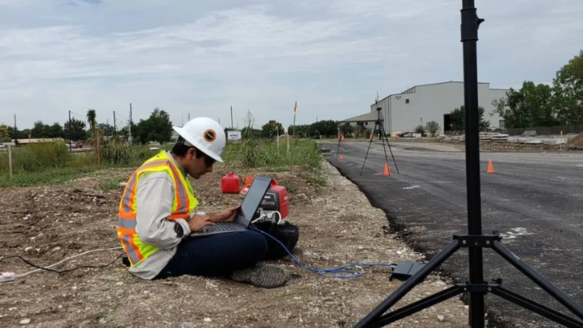 A man sits on the ground and looks at a laptop on an overcast day. He sits on the grass, next to a road, with some equipment set up in the foreground and the background.