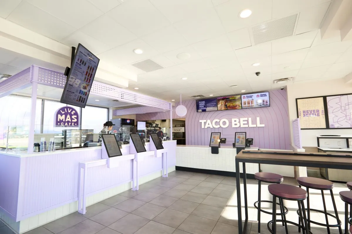 The front counter of a Taco Bell Live Mas Cafe with various kiosks and stools and tables for seating