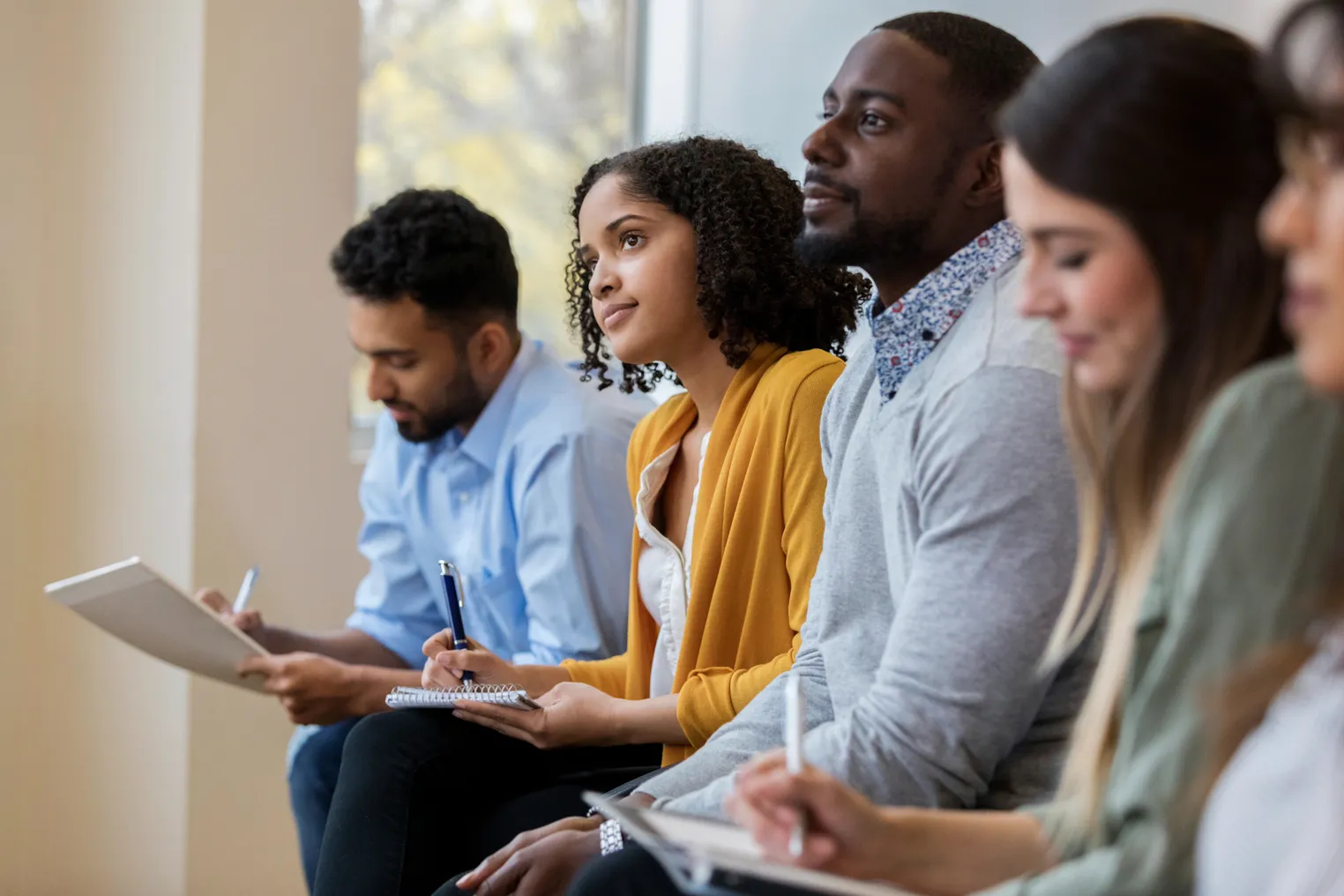 A group of business people sit in a row in a training class. They look at an unseen speaker as they concentrate on his lecture.