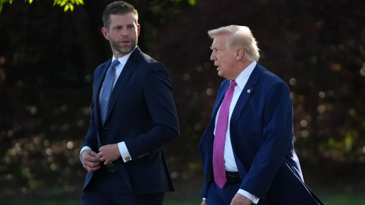 Two people in three-piece suits walking on grass and talking.