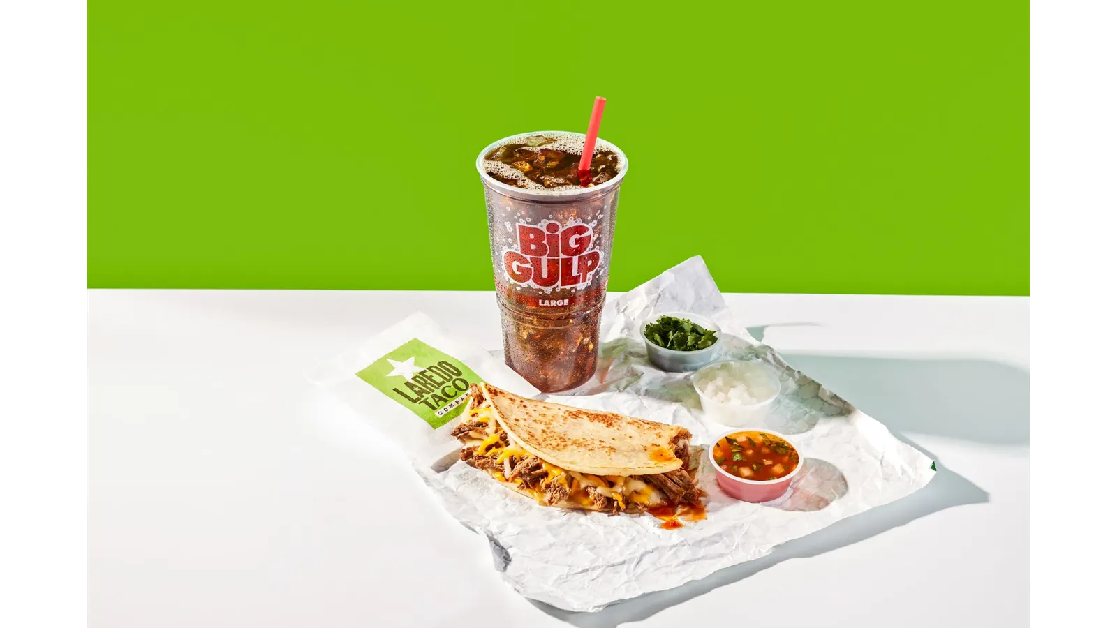 A photo of a taco with toppings and a large cup of soda. The cup says Big Gulp and the wrapper under the taco says Laredo Taco.
