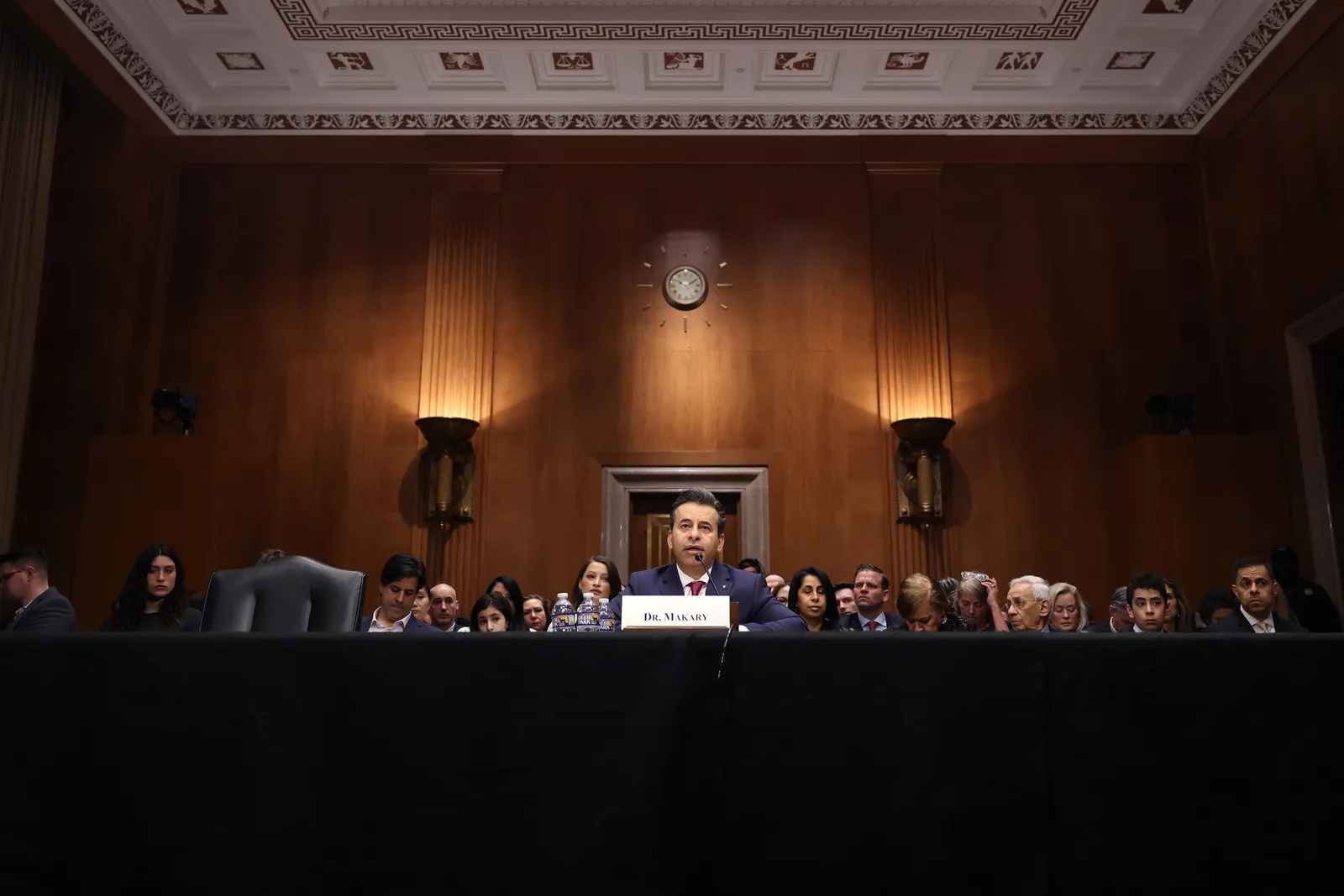 A man sits in the center of a long table with a sign in front of him that reads "Dr. Makary," while a crowd of people sit in several rows behind him in a dark, wood-paneled government building.