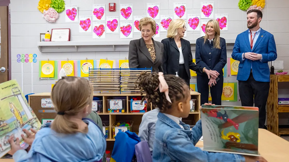 Four adults are standing in the back of a classroom. In the forefront are two students sitting at desks holding books open.