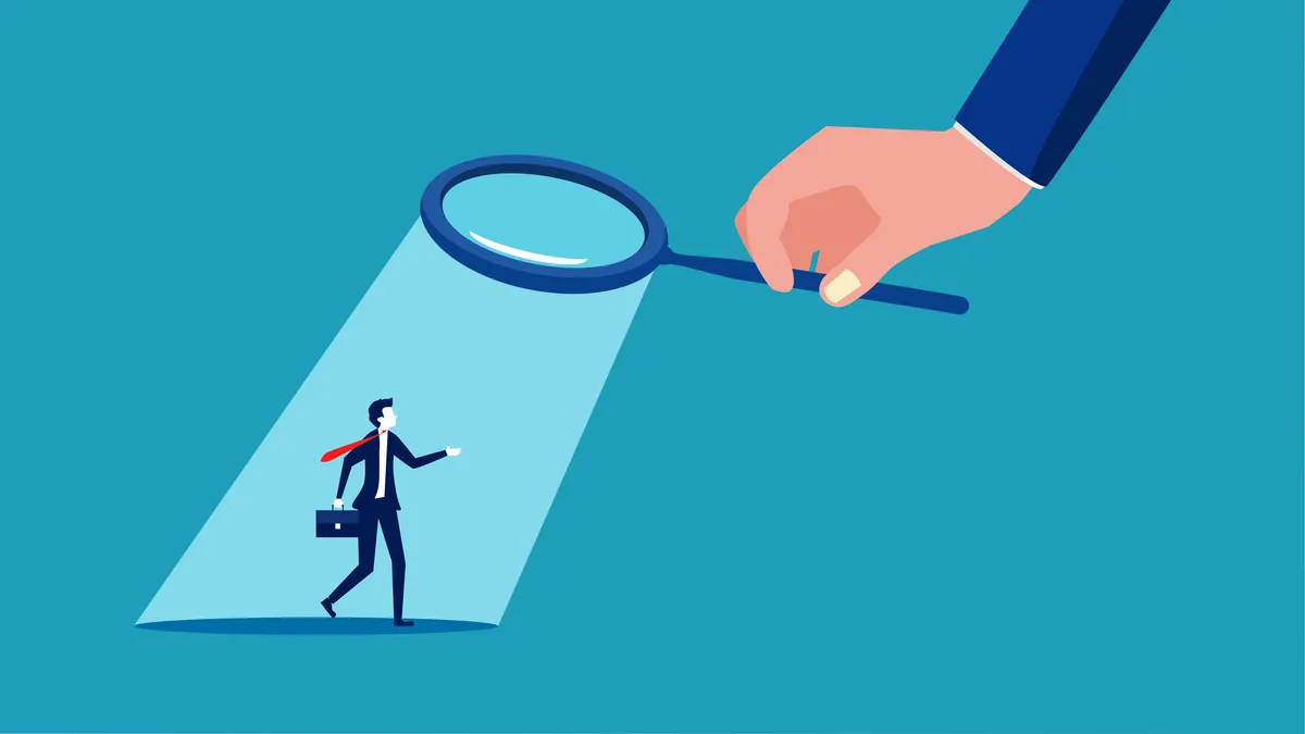 A large hand of a man holds a magnifying glass on a miniature executive, with a spotlight shining on the executive