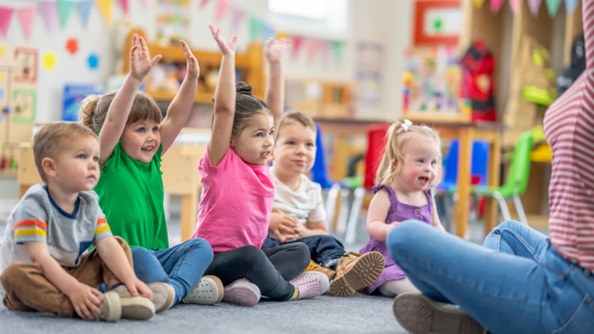 A handful of young children sit on a carpet in a classroom with their hands raised. Part of the body of an adult can be seen in the far right of the photo.