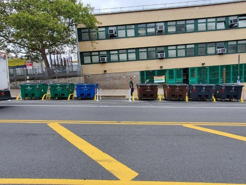 Rows of large waste containers in wheels out front of a New York City school building