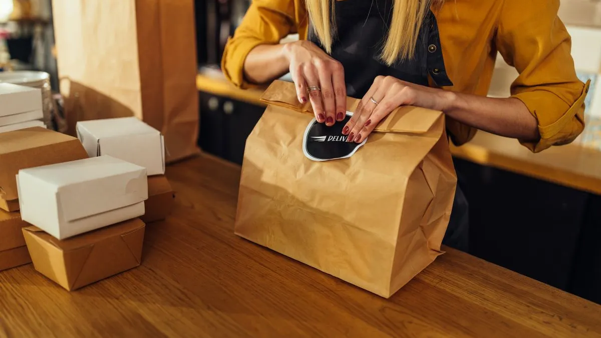 A woman is closing a paper bag with a round black sticker that says delivery. The woman is wearing a yellow shirt and black apron.