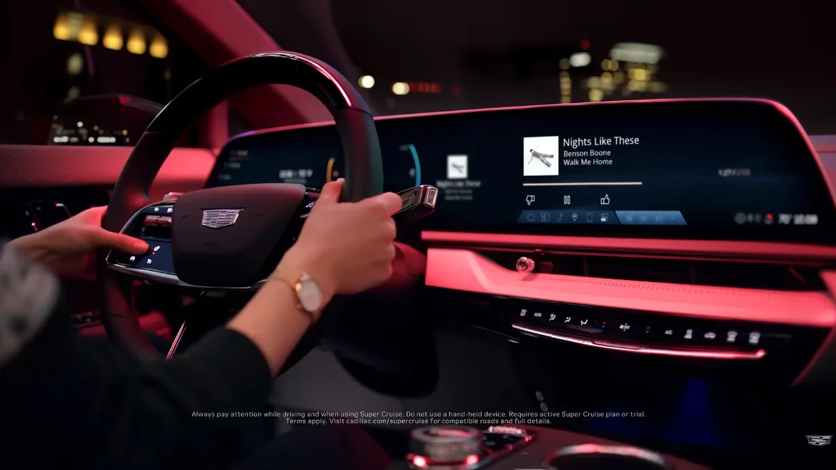 Inside a car, two hands are placed on a steering wheel with a Cadillac logo. Past the steering wheel is an infotainment system and digital cluster, typical of modern cars, showing the song "Nights Like These" by Benson Boone is playing.