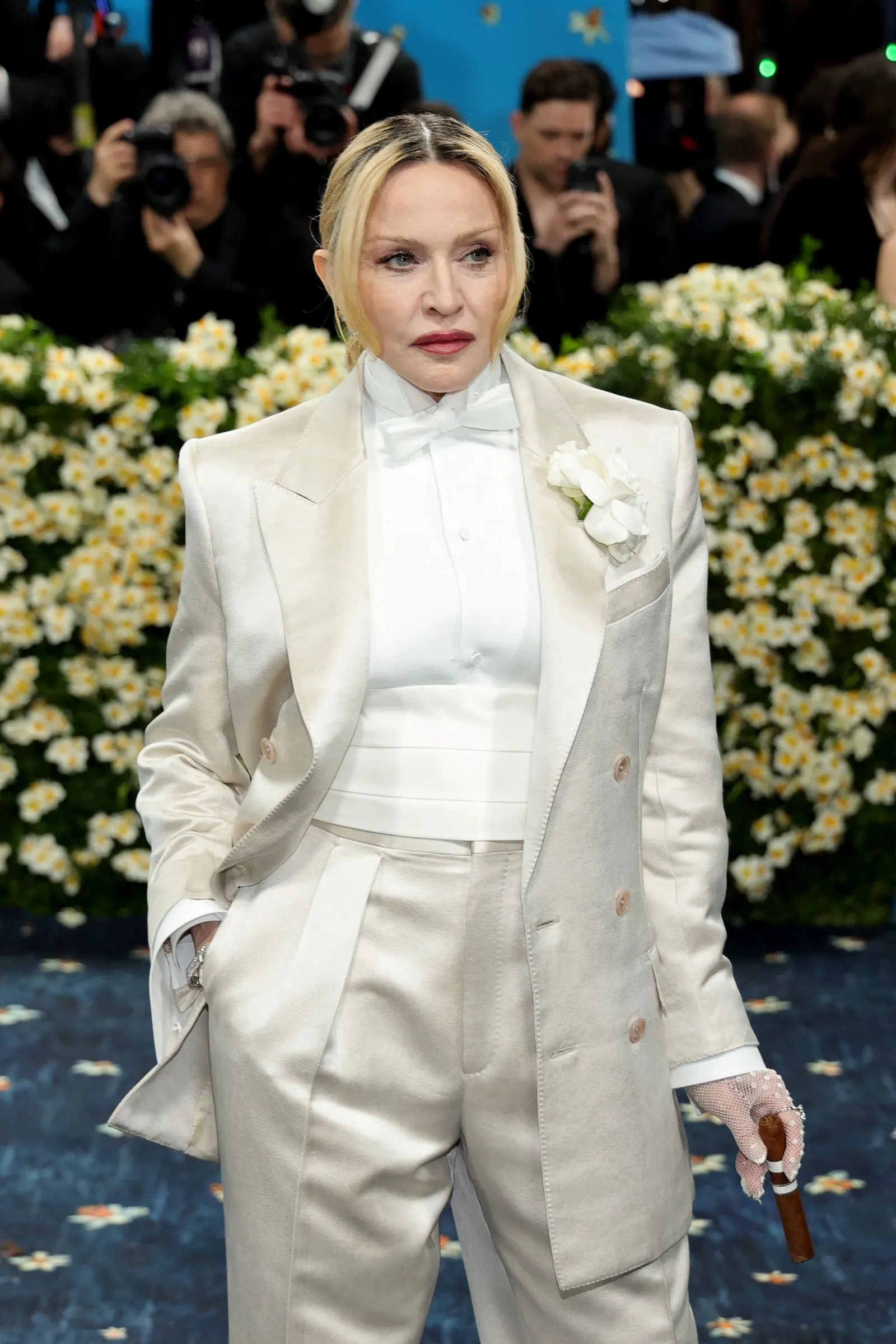 A woman in a white tuxedo stands on a blue carpet holding a cigar.