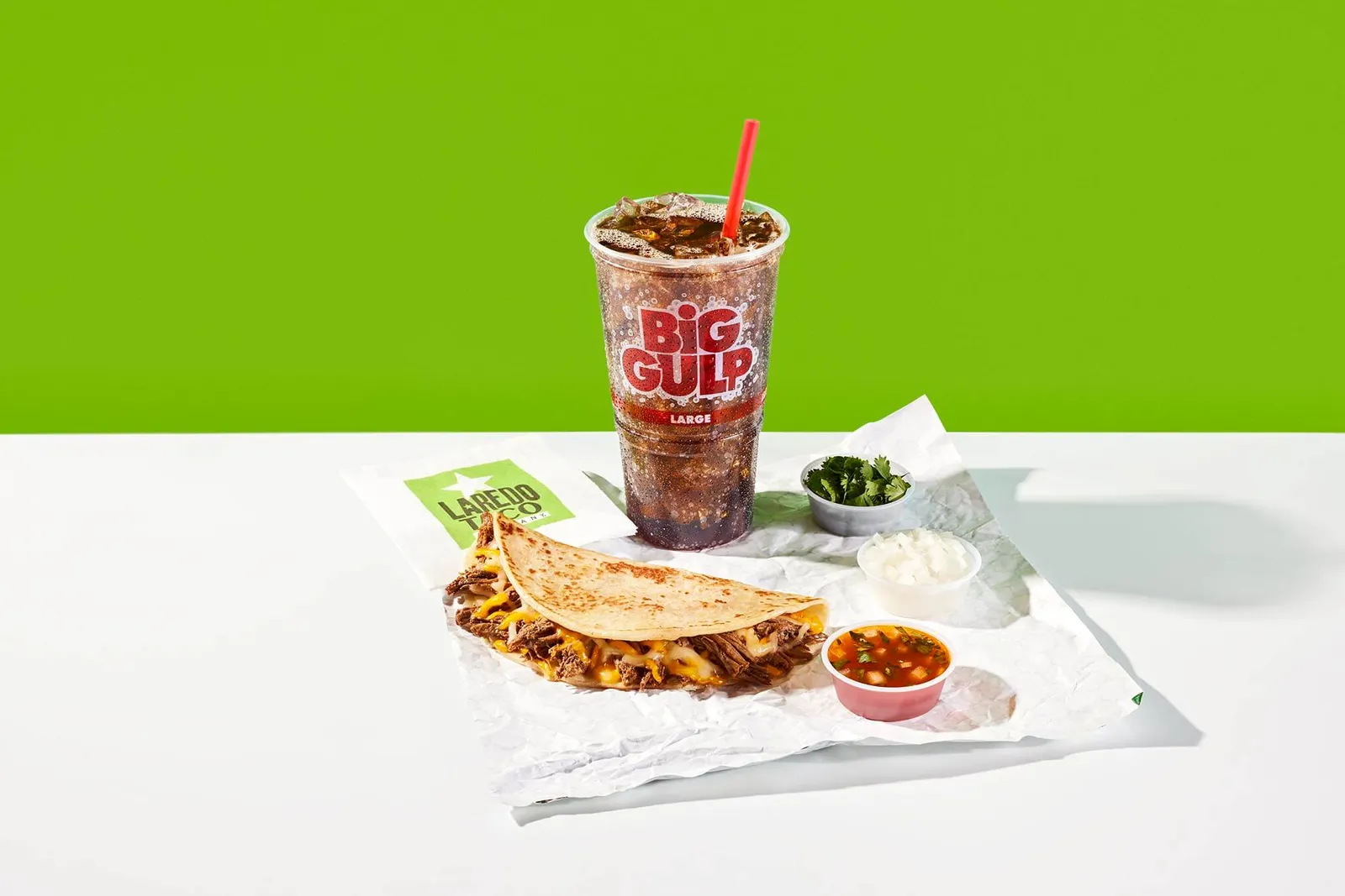 A photo of a tsco on a napkin surrounded by a cup of soda and several cuts of garnishes and dips. the napkin says Laredo Taco and the soda cup says Big Gulp.