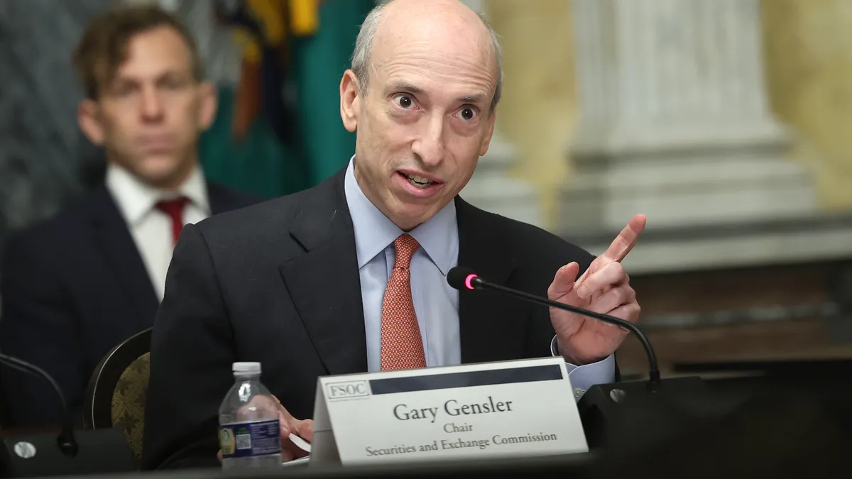 Gary Gensler is in focus speaking with a finger raised in front of a name card. An aide sits behind him out of focus.