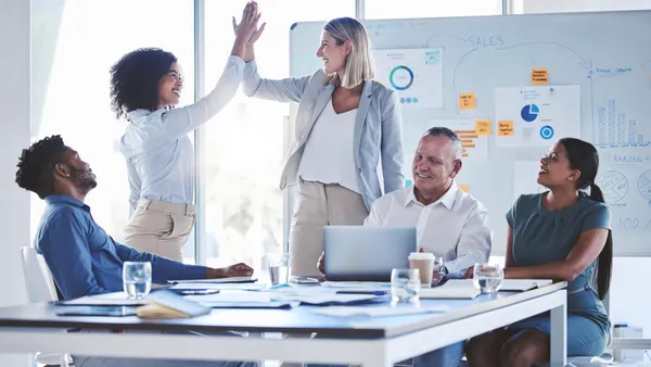 A diverse group of five colleagues in a bright office brainstorm. Two women high-five, conveying success and teamwork. Charts and notes are visible on a whiteboard.