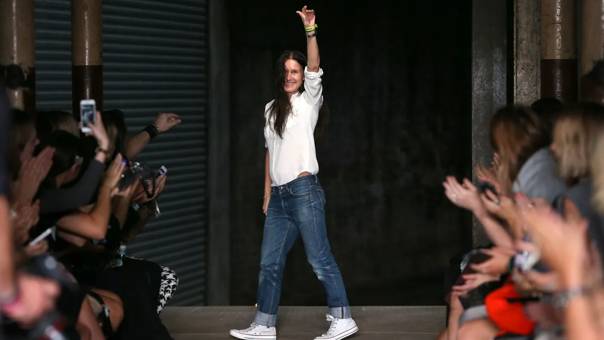 A person in jeans and a white T-shirt waves at the back of a fashion runway as the audience applauds from the sidelines.