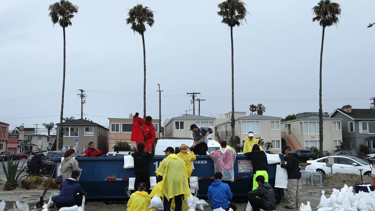 People in rain ponchos stand on a beach surrounded by sandbags. Two people with shovels stand on a large container.