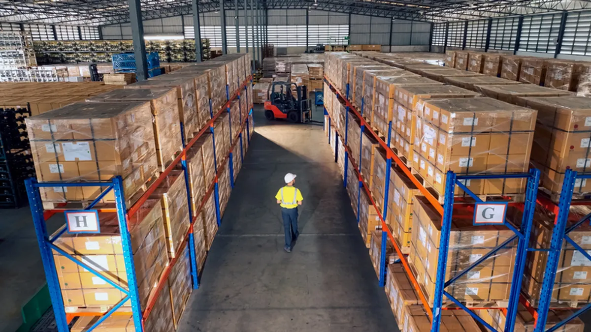 A person in a yellow safety vest walks between tall shelves stacked with boxes in a large warehouse. A forklift operates in the background, conveying efficiency and organization.
