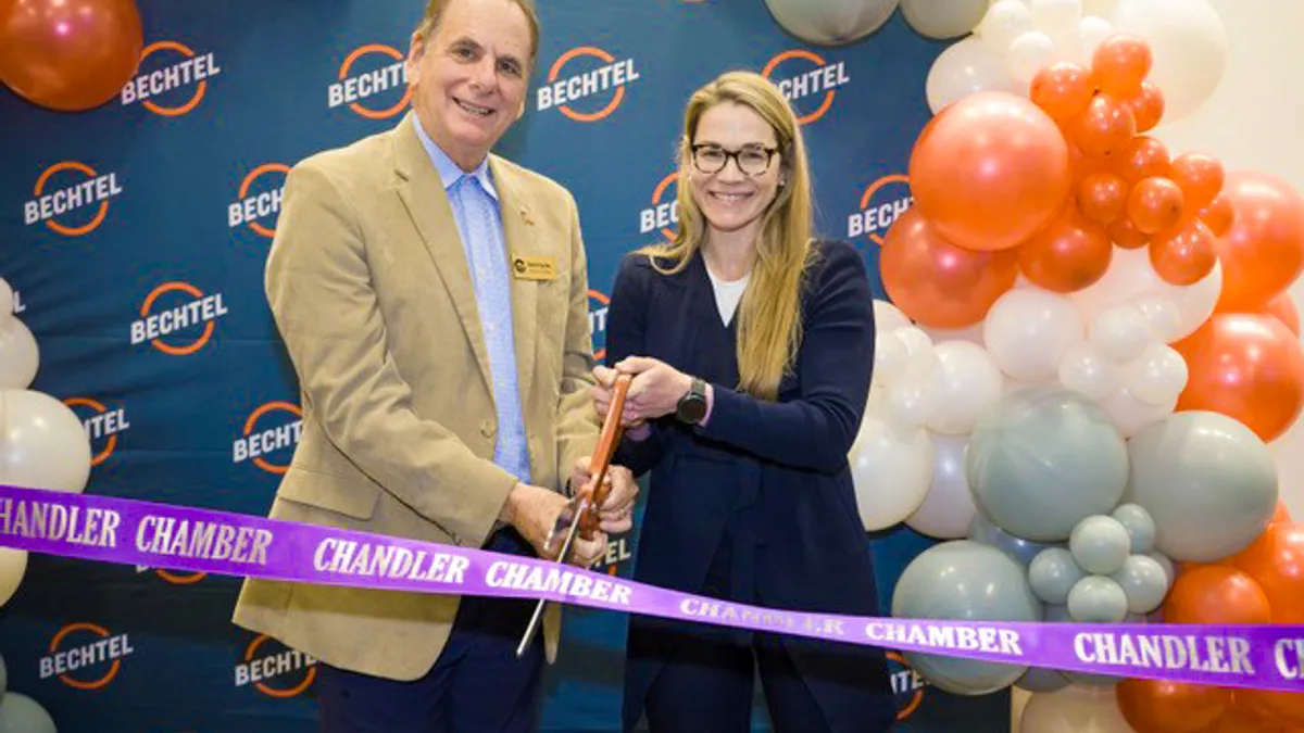 A man and a woman stand in front of a Bechtel-themed backdrop with balloons and ribbons to the side. They're smiling, and standing in front of a ribbon that needs to be cut to open a new office.