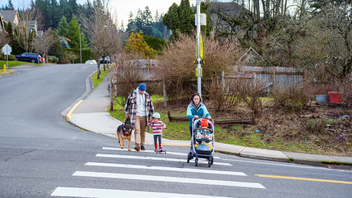 Two adults, a young child and baby in a stroller, accompanied by a dog, cross the street amond trees and homes in the background.
