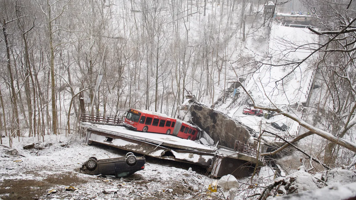 A snow covered bridge is broken in the middle, with a red bus jammed into the crack. A car is on its back on the embankment in front of the bridge.