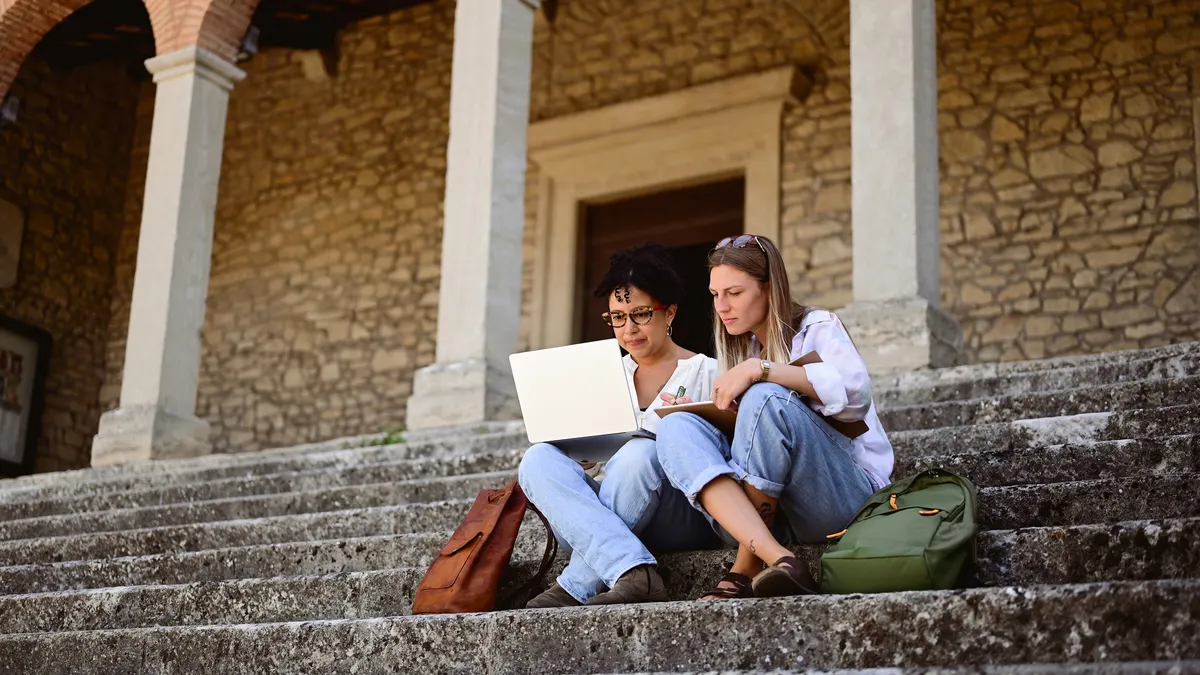 Two people sit on the steps of a college building looking at their laptops.