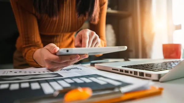 A person in an orange sweater uses a tablet at a desk. Nearby are a laptop, documents with charts, and a pencil. Sunlight filters through a window.