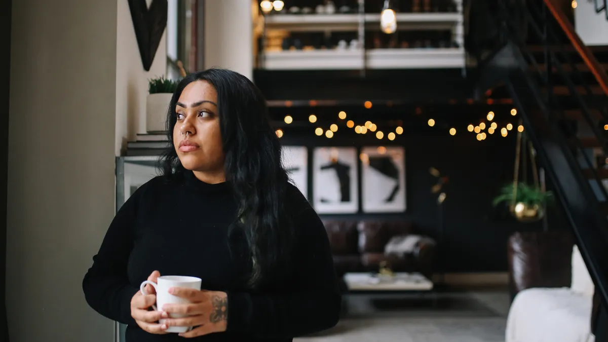 An Indigenous millennial holds a cup of coffee and looks seriously at the camera