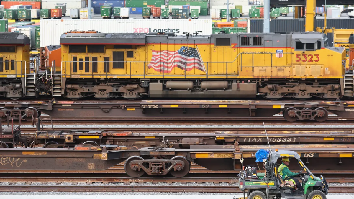 A man in a hardhat drives in a small utility vehicle with a yellow train with American flag in the background