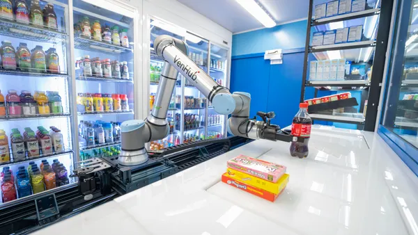 A photo of a robotic, automated c-store. A robotic arm is setting a bottle on a white counter next to three boxes of candy.