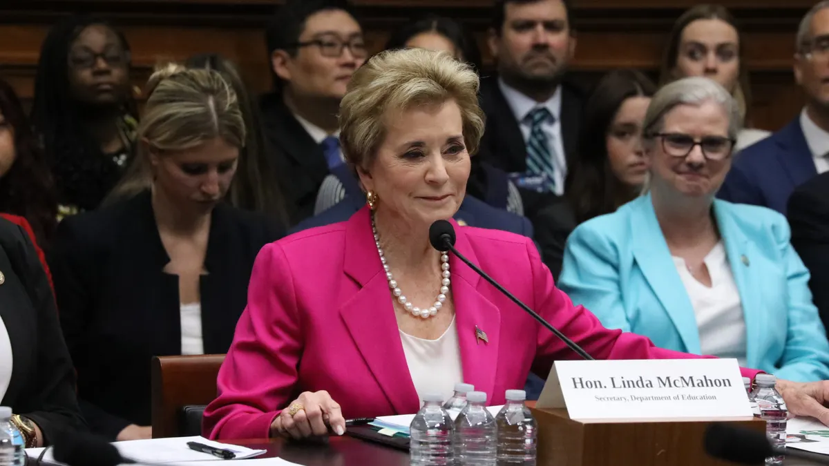 A person sits at a table with a microphone. Water bottles are nearby and people are seated in rows behind the person.