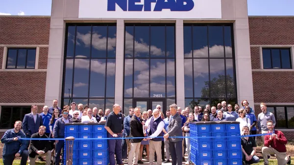 Dozens of people stand behind two people cutting a ribbon in front of a building bearing Nefab's logo.