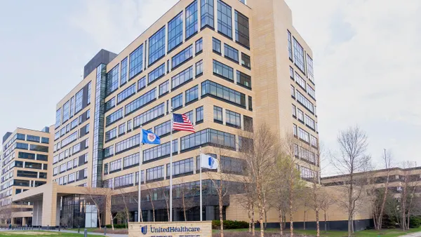 A large modern office complex with tan brick and glass exterior, multiple stories high. In front of the building are three flagpoles flying the U.S. flag, a blue logo flag, and a corporate flag. A sign at the entrance reads “UnitedHealthcare” with a street address, and the surrounding grounds are landscaped with grass and young trees.