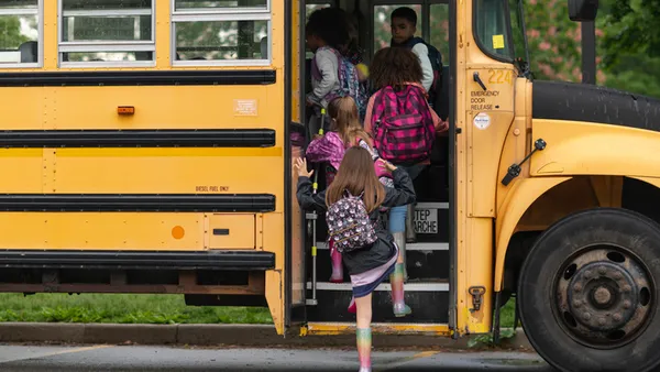 A yellow school bus is parked outside. A handful of students wearing rain jackets and  backpacks are getting on the bus.