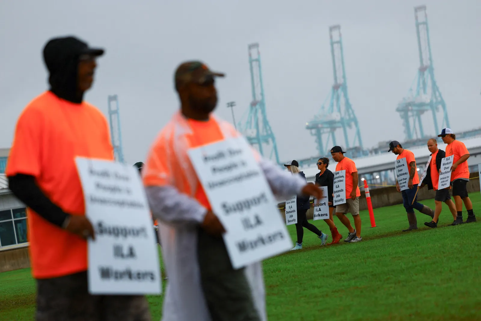 Blurred union members wearing orange shirts and holding signs stand in the forefront, while other protestors are in focus in the background. Several cranes can be seen in the distance.