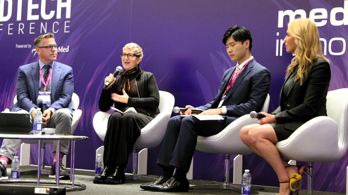 Four people sit side by side on a stage. The second person from the left holds a microphone and speaks.