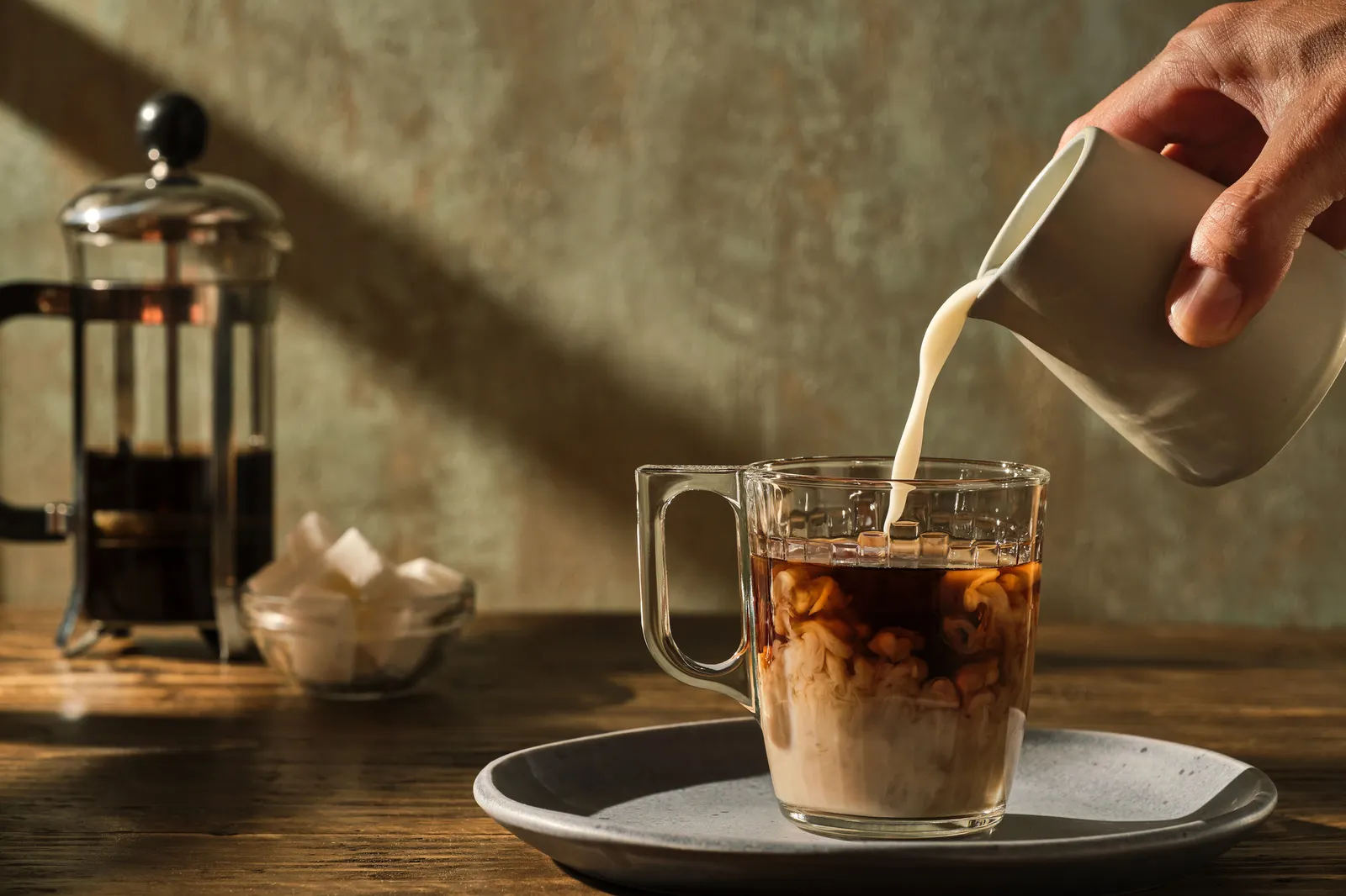 A hand pours cream out of a beige pitcher into a class mug containing coffee sitting on a grey saucer. The coffee is on a wooden table and a French press and sugar bowl are in the background.