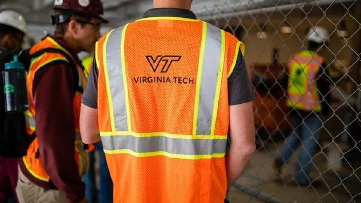 Two people stand with orange safety vests near a chain-link fence. One student has the logo of Virginia Tech on their back.