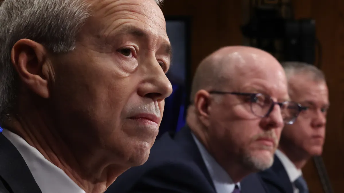 Three people in suits sit on a dias at a Congressional hearing.