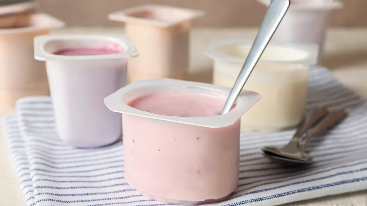 Several white, plastic yogurt cups containing different colored yogurt, one of which has a metal spoon sticking out of it.