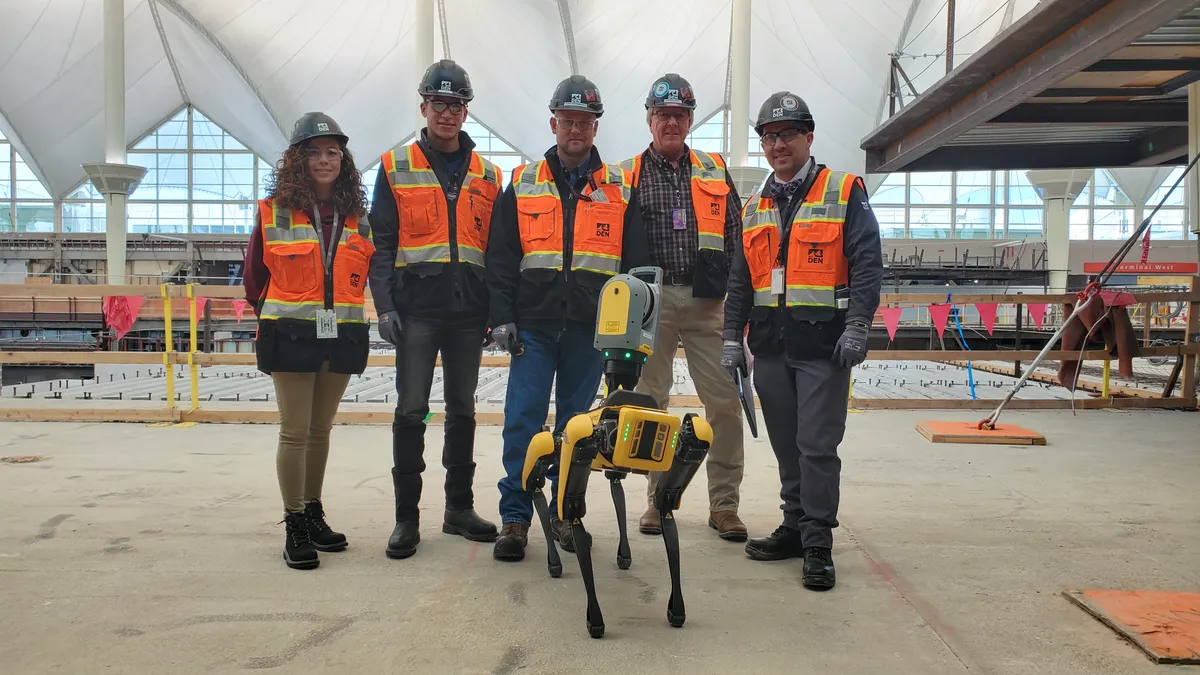 Four workers stand in orange vests and white hardhats on a construction site. In front of them is a robot that stands quadripedal like a dog. The group is posed for the photo, shoulder-to-shoulder.