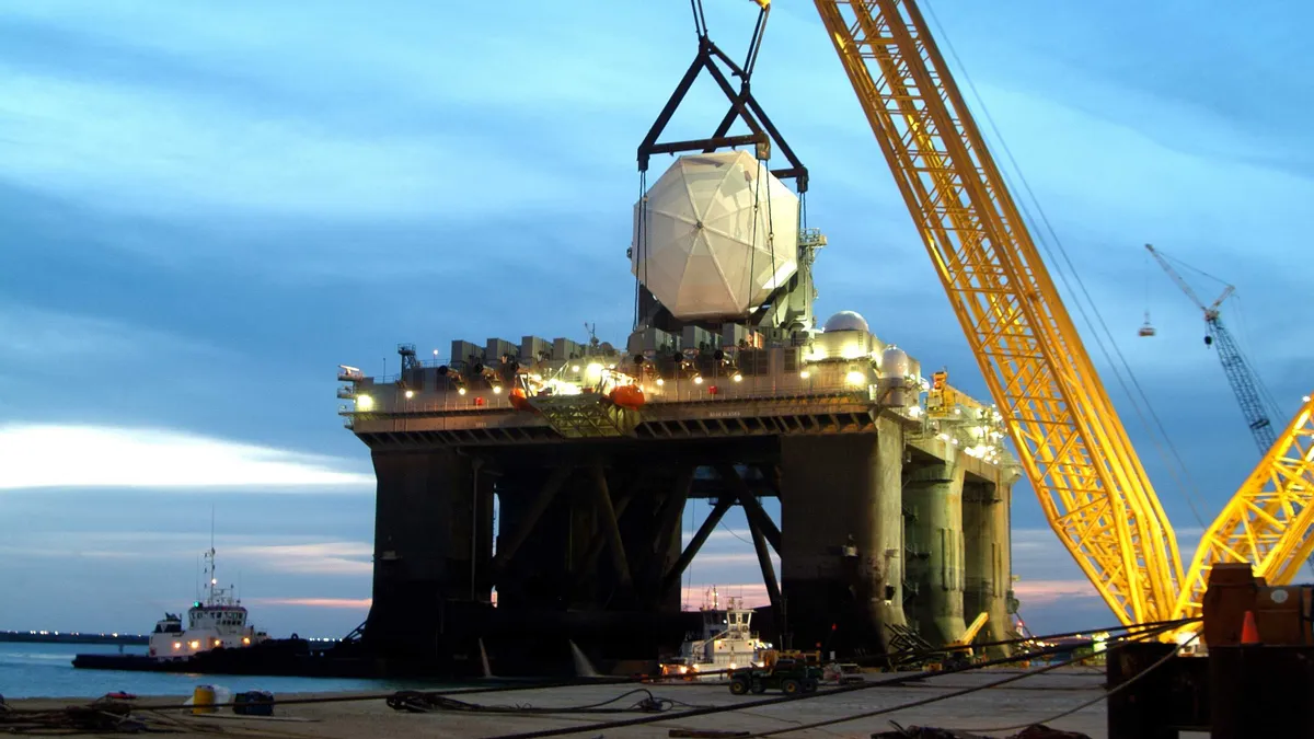 A massive yellow crane hoists a massive object above the water in front of an oil rig.