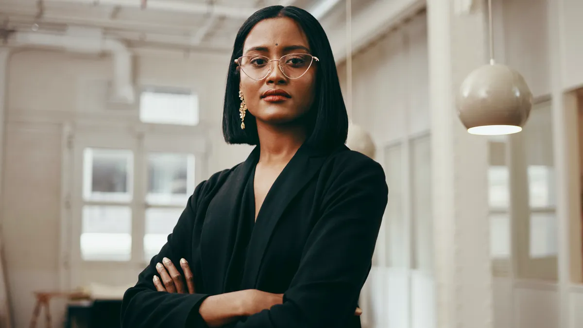 A South Asian person with a bindi stands with their arms folded for a portrait