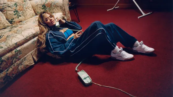 A person laying against a couch on the floor in a tracksuit and sneakers with a phone pressed to their face.