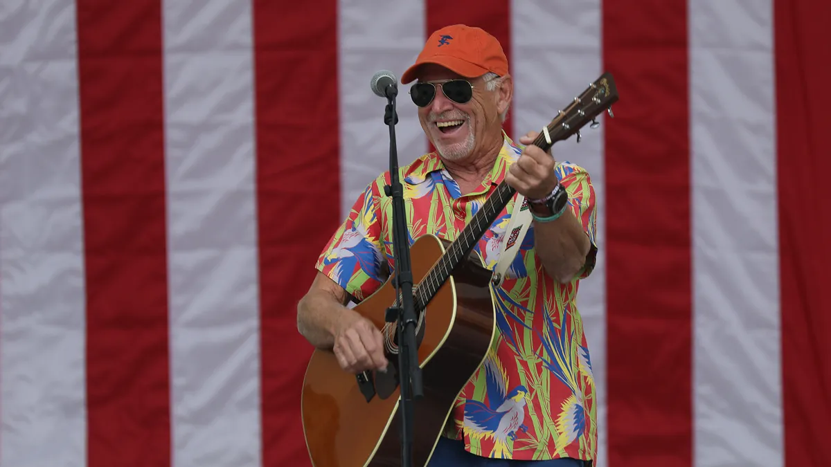 Jimmy Buffett wears a yellow Hawaiian shirt and holds an acoustic guitar in front of a large American flag.