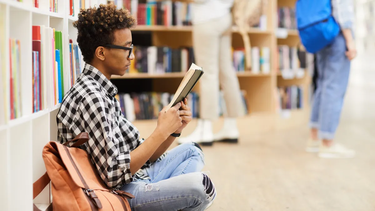 A serious teenager sits with crossed legs on a library floor, reading an interesting book.