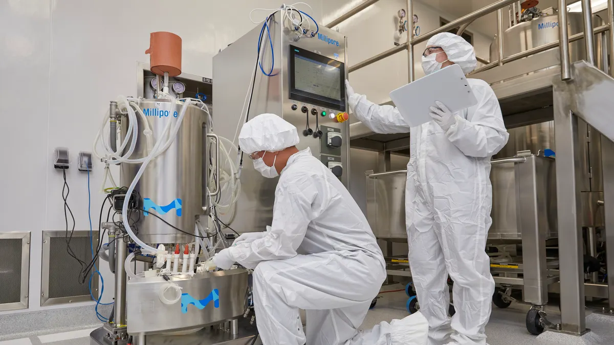 Two people in laboratory gowns inspect biotechnology equipment with lettering spelling Millipore.