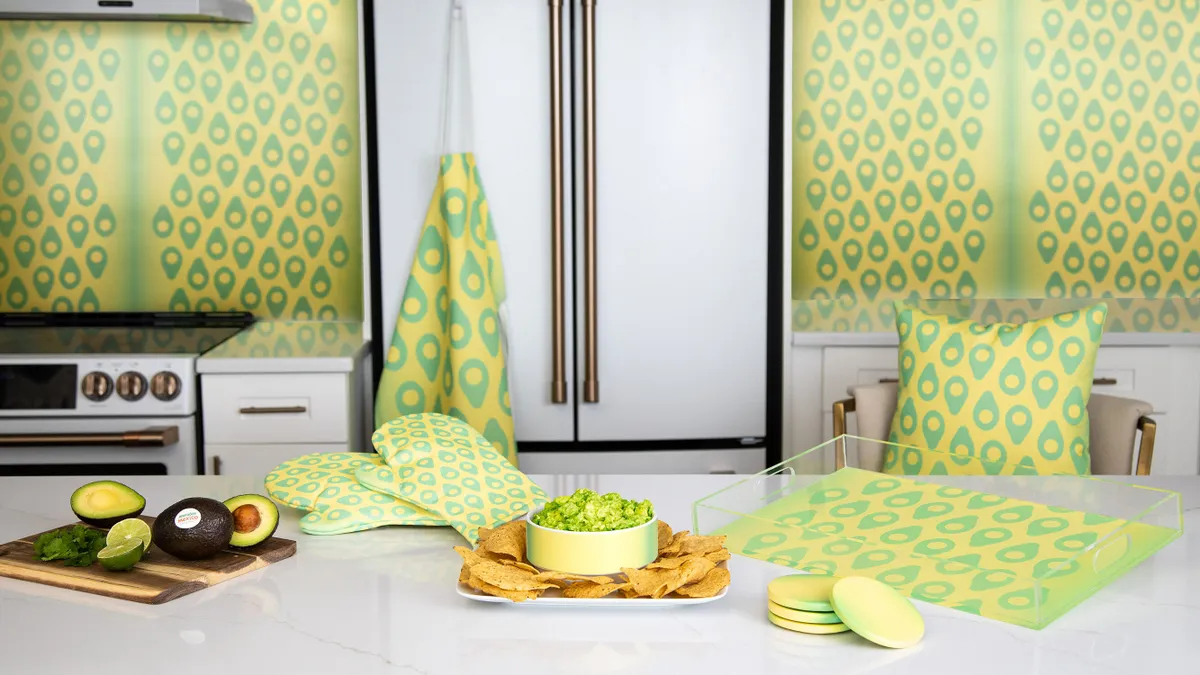 A white marble kitchen countertop displays, from left to right, an avocado and limes on a cutting board, oven mitts, chips and guacamole and coasters and wall paper themed around a new Pantone color.