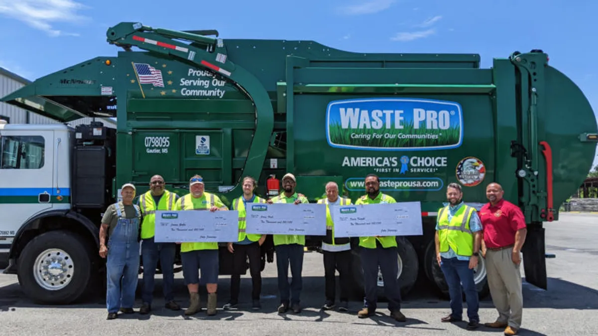 Workers in yellow uniforms/vests hold oversized checks while standing in front of a green trash truck